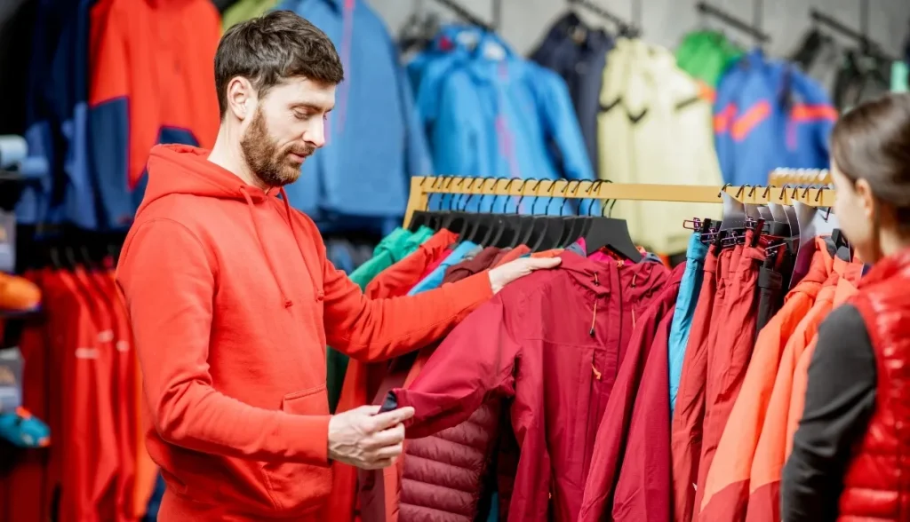 A man with a beard wearing a red hoodie is examining a red jacket on a clothing rack in an outdoor apparel store.