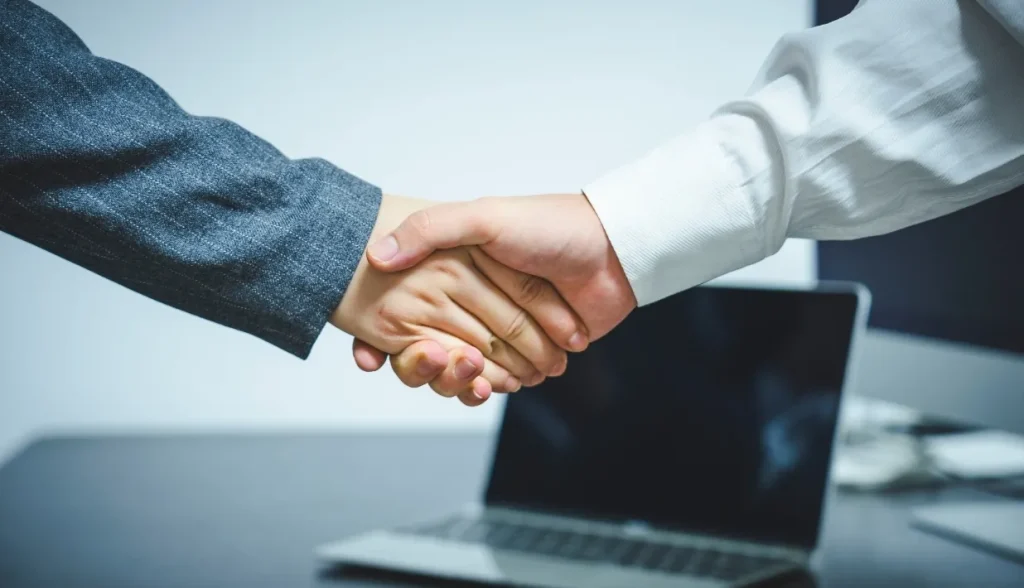 Two businesspeople shaking hands over a laptop in an office setting.