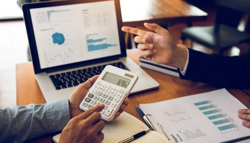 Two people in a business meeting: one holds a calculator, the other points at a laptop showing data charts, with financial documents on the table.