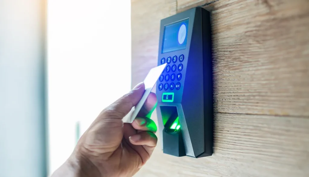 Mano acercando una tarjeta de proximidad de PVC en blanco a un lector multifunción de control de acceso RFID con teclado, sensor de huellas dactilares y teclas numéricas retroiluminadas, instalado en una pared de madera.