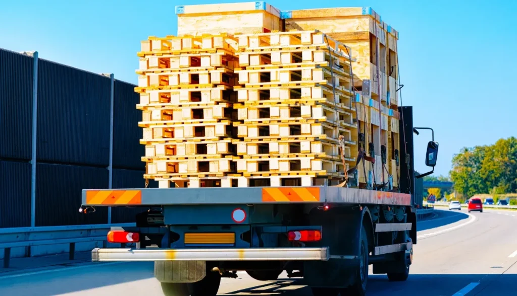 Flatbed highway truck loaded with stacks of empty wooden pallets in transit, long-distance transport managed via RFID pallet tracking.