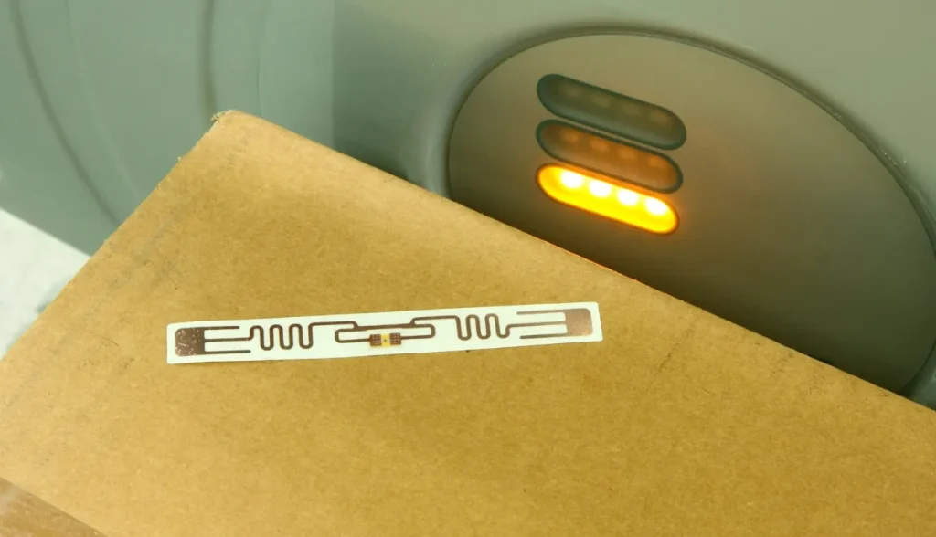 Etiqueta adhesiva alargada de RFID UHF pasiva fijada en una caja de cartón marrón junto a la luz indicadora LED del lector RFID; hardware de escaneo para la verificación y el control de activos en la cadena de suministro.