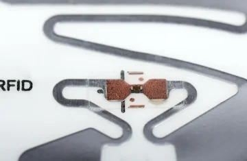UHF RFID Chip Comparison Performance Features and Price Macro close-up of UHF RFID inlay tag with copper antenna and tiny RFID microchip, printed "RFID" text on white substrate, ultra-high frequency radio frequency identification sticker hardware.