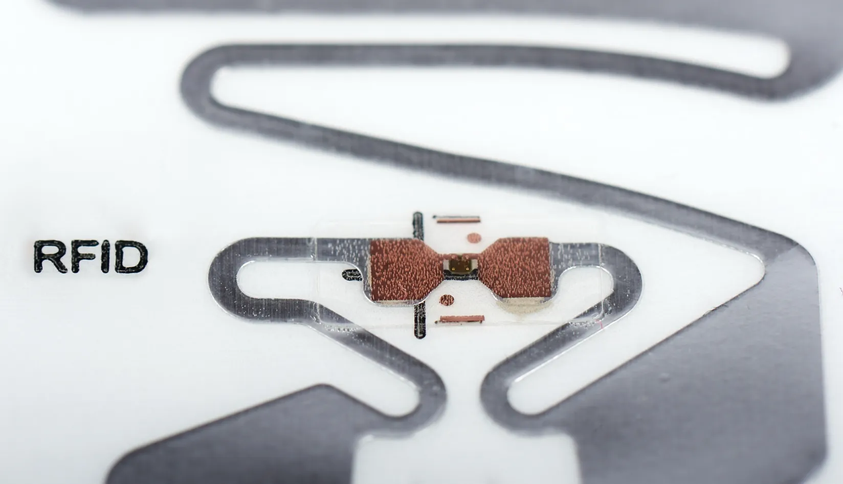 UHF RFID Chip Comparison Performance Features and Price Macro close-up of UHF RFID inlay tag with copper antenna and tiny RFID microchip, printed "RFID" text on white substrate, ultra-high frequency radio frequency identification sticker hardware.